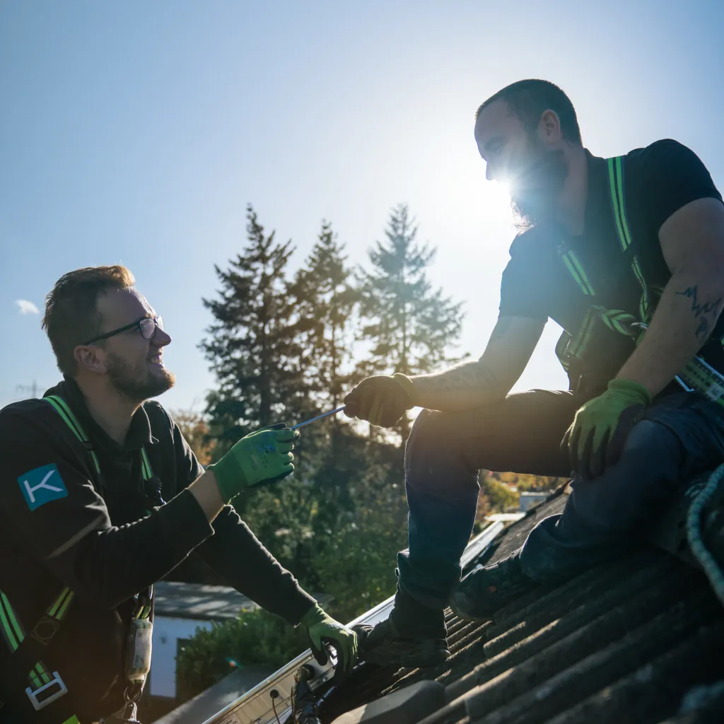 Zwei Mitarbeiter von Mike Kolbe Heizungstechnik bei der Arbeit auf einem Hausdach. Ein Mitarbeiter reicht seinem Kollegen Werkzeug hoch.